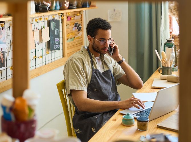 Young male wearing a smock sitting at a desk in front of a computer while talking on the phone